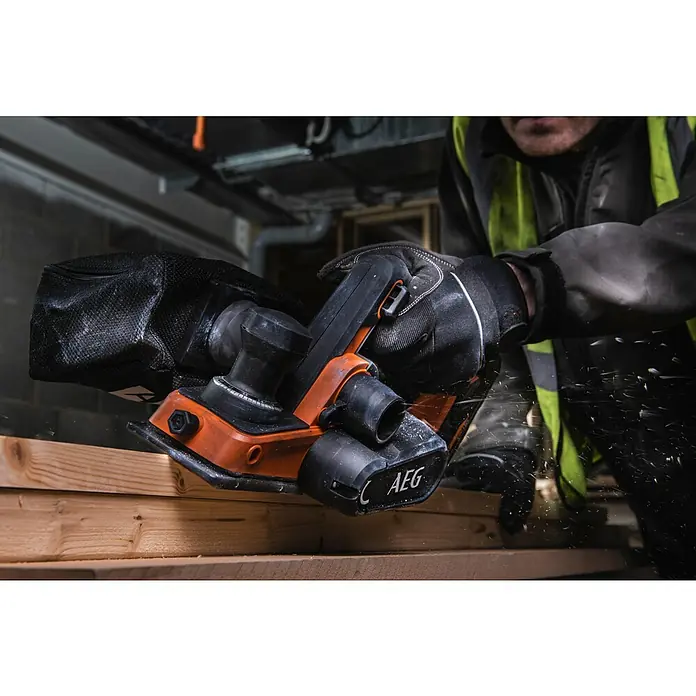 AEG Akku-Hobel BHO18BL-0 - 18 V, Ohne Akku, Hobeltiefe: 0 mm - 2 mm, Leerlaufdrehzahl: 14.300 U/min - Eine Person hobelt mit einem orange-schwarzen AEG Elektrohobel Holz, Holzspäne fliegen. Ein Staubbeutel ist angebracht.