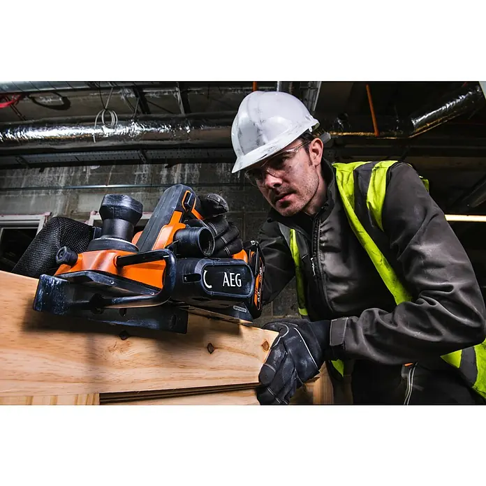 AEG Akku-Hobel BHO18BL-0 - 18 V, Ohne Akku, Hobeltiefe: 0 mm - 2 mm, Leerlaufdrehzahl: 14.300 U/min - Mann in Schutzkleidung bedient einen orange-schwarzen AEG Elektrohobel/Schleifer auf einem Holzbrett.
