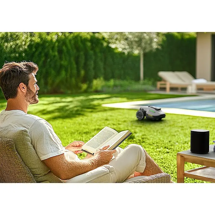 Mammotion Mähroboter Yuka Mini 800800 m², Schnittbreite: 190 mm, Satellitengestützte Navigation, RTK - Ein Mann entspannt sich mit einem Buch, während der Rasenmähroboter im Garten arbeitet, und genießt kabellose Musik auf einem Smart Speaker.