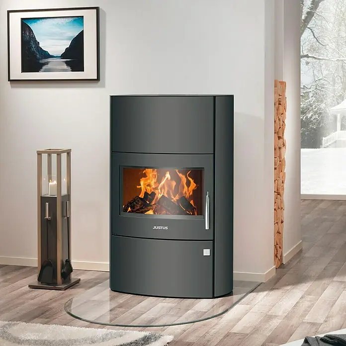 Justus Kaminofen Agero W+ - 7 kW, Raumheizvermögen: 144 m³, Material Verkleidung: Stahl, Schwarz - Ein Justus Kaminofen mit Glasplatte und brennendem Holz, umgeben von dekorativem Zubehör und einem Holzstapel.
