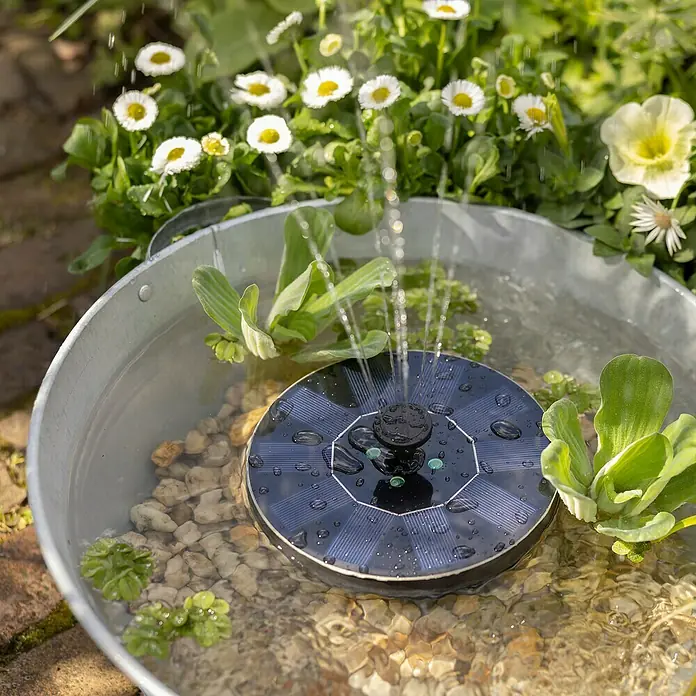 Fuente de jardín LED Lioni - Plástico, Negro - Una fuente solar flota en una tina de zinc rodeada de plantas y rocas, creando un suave elemento acuático en el jardín.