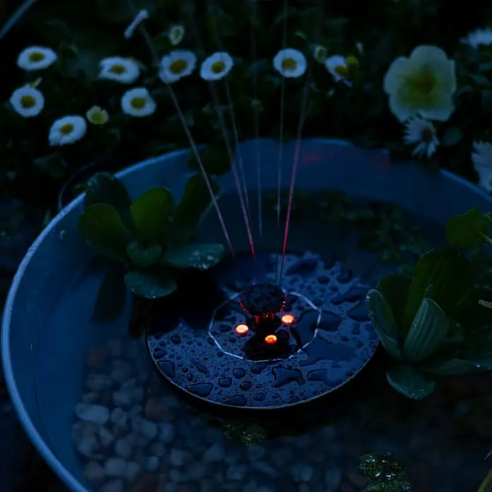 Fuente de jardín LED Lioni - Plástico, Negro - Primer plano de una bomba de fuente solar en agua, rodeada de flores y plantas.