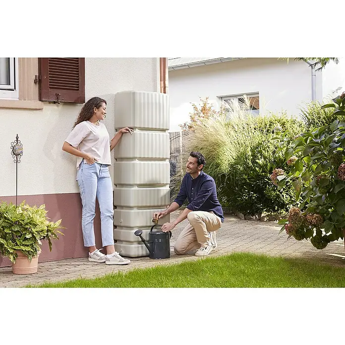 4rain Regenspeicher Slim - 350 l, Sandbeige, 1 Stk. - Eine stilvolle Regenwassertonne mit einer Frau, die sich anlehnt, und einem Mann, der Wasser in eine Gießkanne abfüllt, im Garten.