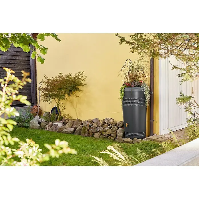 4rain Regenspeicher Milo - 230 l, Graphitgrau, 1 Stk. - Grauer Regenspeicher mit Bepflanzung auf einer grünen Rasenfläche vor gelber Wand. Ein dekorativer Hingucker für den Garten.