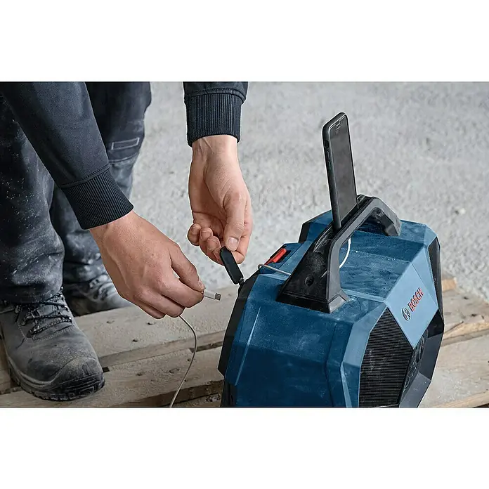 Bosch Professional AMPShare 18V Bluetooth-Lautsprecher GPB 18V-6 C - 18 V, Ohne Akku - Person steckt Kabel in blaues Bosch Baustellenradio mit Smartphone im Halter auf der Baustelle.