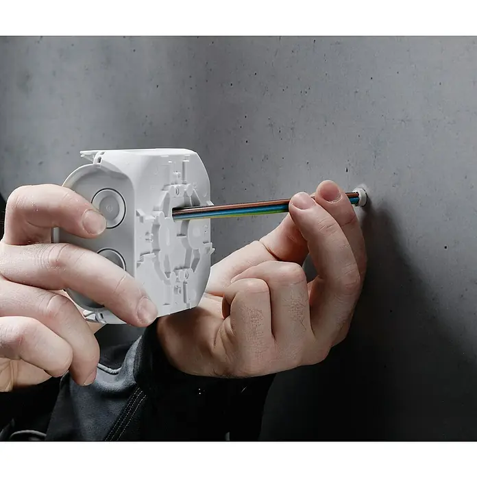 Hensel  Abzweigkasten Kabel DK0600G - 130 x 130 x 77 mm - Ein Installateur führt ein Elektrokabel durch eine weiße Abzweigdose in eine graue Betonwand ein.