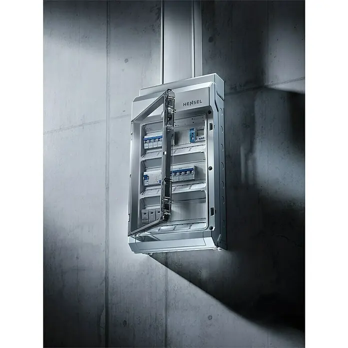 Hensel  Aufputz-Verteiler KV9224 - Anzahl Reihen: 2 Stk., 29,5 x 45,8 cm, Grau - Hensel-Verteilerkasten an einer Betonwand montiert, die transparente Tür geöffnet, zeigt die internen Sicherungen und Schalter für die elektrische Installation.