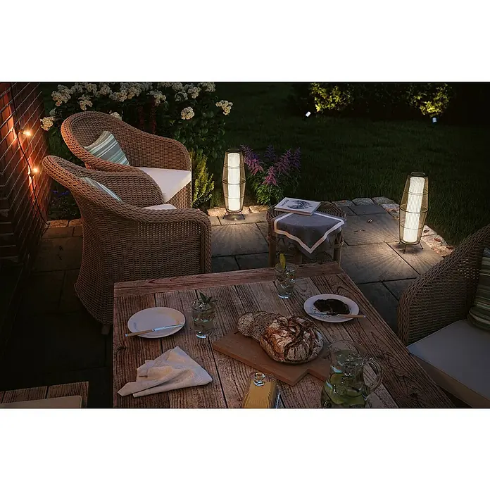 Paulmann Plug & Shine LED-Außenleuchte Basket - 8 W, Farbe: Braun, IP44, 20 x 60 cm - Ein gemütlicher Außenbereich mit zwei Korbsesseln, einem Holztisch mit Speisen und Getränken, beleuchtet von dekorativen Gartenleuchten.