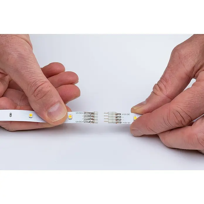 Paulmann Tira LED Eco cálido - Largo: 5 m, Blanco cálido, 12 W, 750 lm, 12 V - Conexión de tira LED: Primer plano de manos conectando una tira de luz LED con un conector de cuatro pines para una conexión segura de 12V DC.