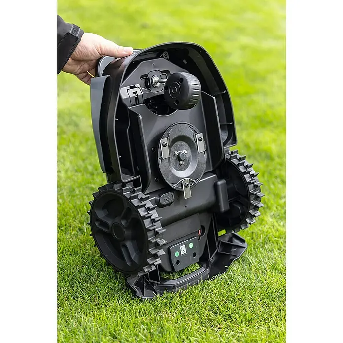 Scheppach Mähroboter BRMR300 - 300 m², Schnittbreite: 16 cm, Induktionsgestützte Navigation, Begrenzungskabel - Nahaufnahme der Unterseite eines Rasenmähroboters mit sichtbaren Rädern und Messern, der auf einer Rasenfläche steht.