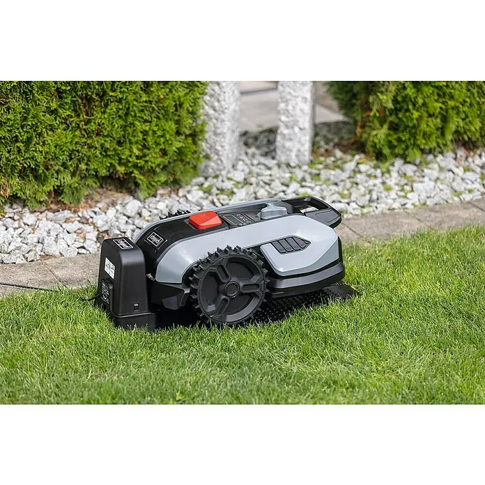 Scheppach Mähroboter BRMR300 - 300 m², Schnittbreite: 16 cm, Induktionsgestützte Navigation, Begrenzungskabel - Der Mähroboter steht auf dem Rasen. Der Roboter ist grau, schwarz und hat einen roten Knopf. Im Hintergrund sind Steine und Büsche.