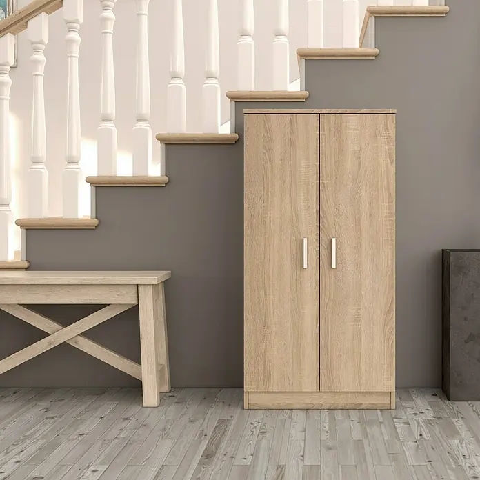Zapatero Basic - L x An x Al: 36 x 55 x 108 cm, Roble Canadian - Un armario estrecho de madera con dos puertas se encuentra debajo de una escalera, proporcionando espacio de almacenamiento adicional en la habitación.