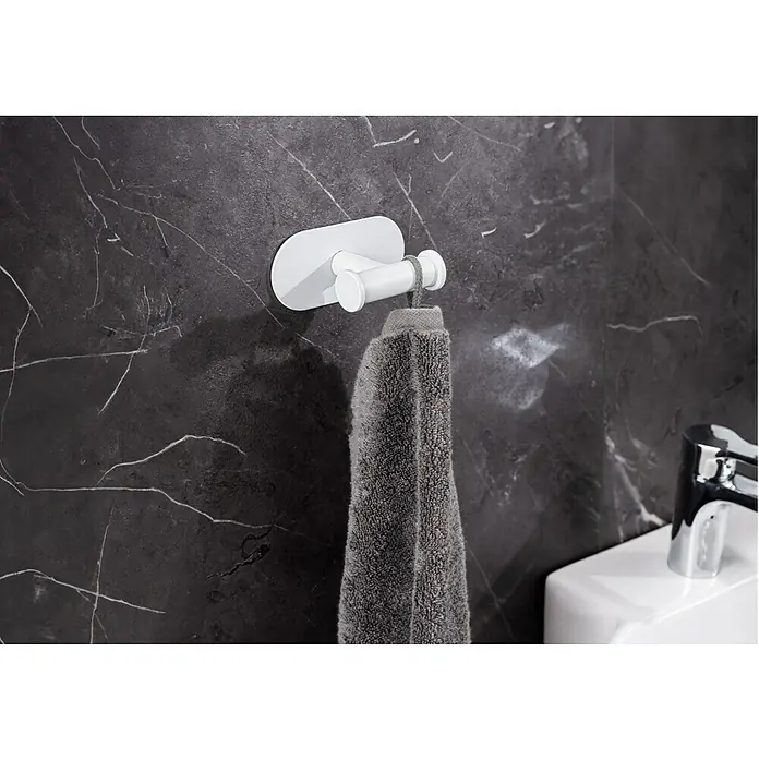 Wenko Orea Colgador de baño - Blanco - Un toallero blanco con una toalla gris cuelga en una pared de mármol oscuro en un baño. El diseño moderno complementa la decoración.