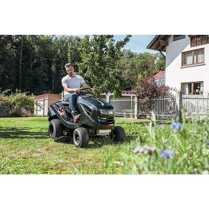 AL-KO Easy Rasentraktor 15-93.2 HDS-A - 452 cm³, Schnittbreite: 93 cm - Ein Mann fährt einen schwarzen AL-KO Aufsitzmäher auf einem grünen Rasen, mit einem Haus und Bäumen im Hintergrund.