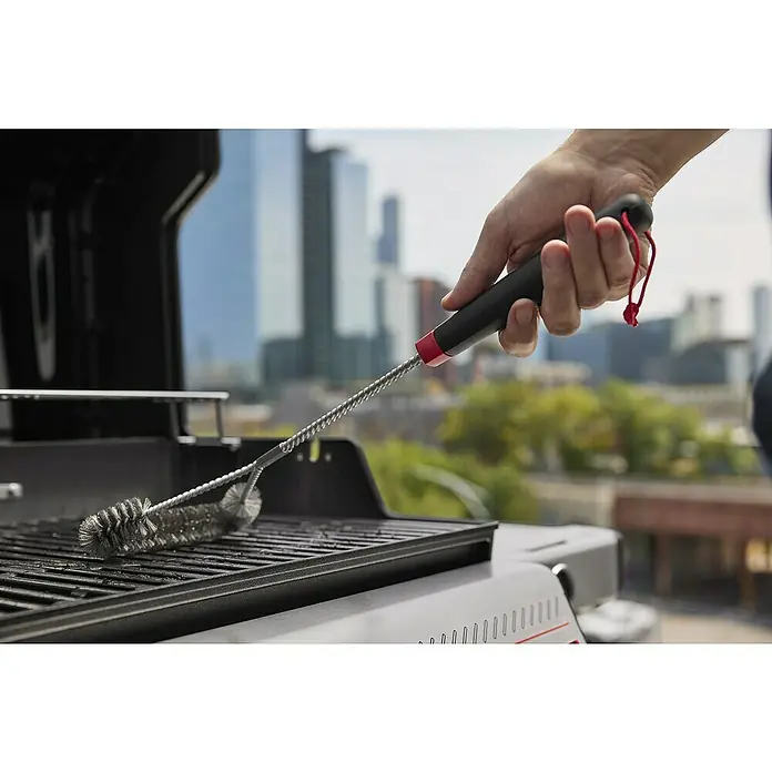 Weber Gasgrill Spirit E-335 GBS - Grillfläche (B x T): 52 x 44 cm, 9,1 kW, Anzahl Brenner: 3 Stk. - Eine Grillbürste mit langem Griff wird verwendet, um einen Grillrost zu reinigen, wobei im Hintergrund eine Stadtansicht zu sehen ist.