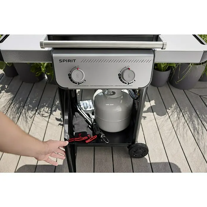 Weber Gasgrill Spirit E-210 - Grillfläche (B x T): 52 x 44 cm, 7,8 kW, Anzahl Brenner: 2 Stk. - Nahaufnahme eines Weber Spirit Gasgrills mit geöffneter Tür, der eine Gasflasche, Grillutensilien und Handschuhe zeigt.