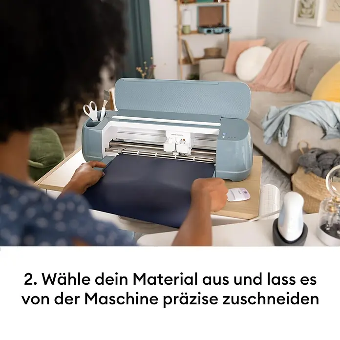 Cricut Maker Schneideplotter-Set Maker 4 + Materials Starter Bundle - 2 -tlg., Bluetooth, Schnittbreite: 29,7 cm, Schnittlänge: 3,6 m - Eine Person führt dunkles Material in eine blau-graue Cricut Maker 4 Schneidemaschine zum präzisen Zuschnitt ein.