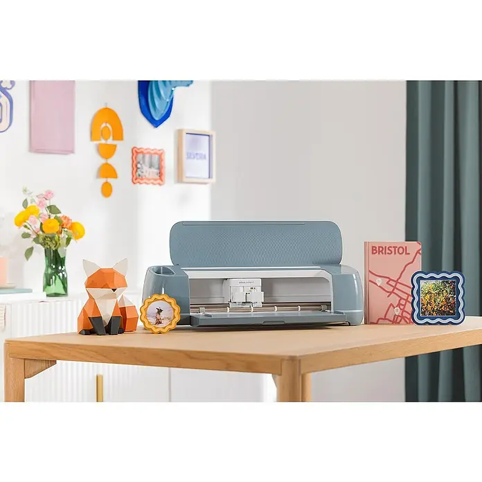 Cricut Maker Schneideplotter-Set Maker 4 + Materials Starter Bundle - 2 -tlg., Bluetooth, Schnittbreite: 29,7 cm, Schnittlänge: 3,6 m - Produktfoto eines Cricut Maker 3 Schneideplotters auf einem Holztisch, mit Bastelarbeiten wie einem Fuchs und Bilderrahmen dekoriert.