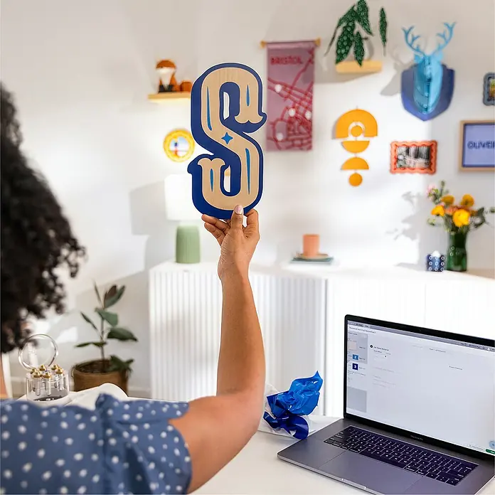 Cricut Maker Schneideplotter-Set Maker 4 + Materials Starter Bundle - 2 -tlg., Bluetooth, Schnittbreite: 29,7 cm, Schnittlänge: 3,6 m - Eine Person hält einen Holzbuchstaben S mit blauen Akzenten vor einem Schreibtisch mit Laptop.