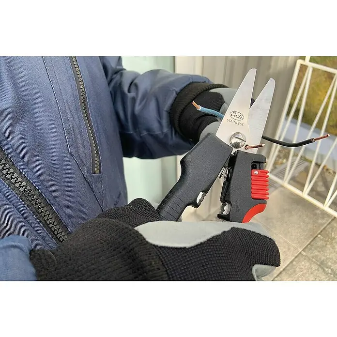 Bessey Tijera universal multiusos Combinox - Largo: 190 mm, Longitud de corte: 40 mm, Acero inoxidable, Negro/Rojo - Primer plano de una persona usando unas tijeras de cable Erdi aisladas para cortar un cable, usadas con guantes de trabajo sobre un fondo borroso.