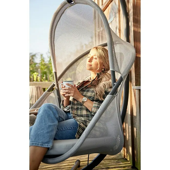 Siena Garden Hängesessel Yobaya mit Schutzhülle - L x B x H: 106 x 123 x 195 cm, Grau - Frau entspannt in einem grauen Hängesessel mit Netzstruktur und genießt eine Tasse Kaffee. Das moderne Design des Sessels fügt sich nahtlos in die Natur ein.