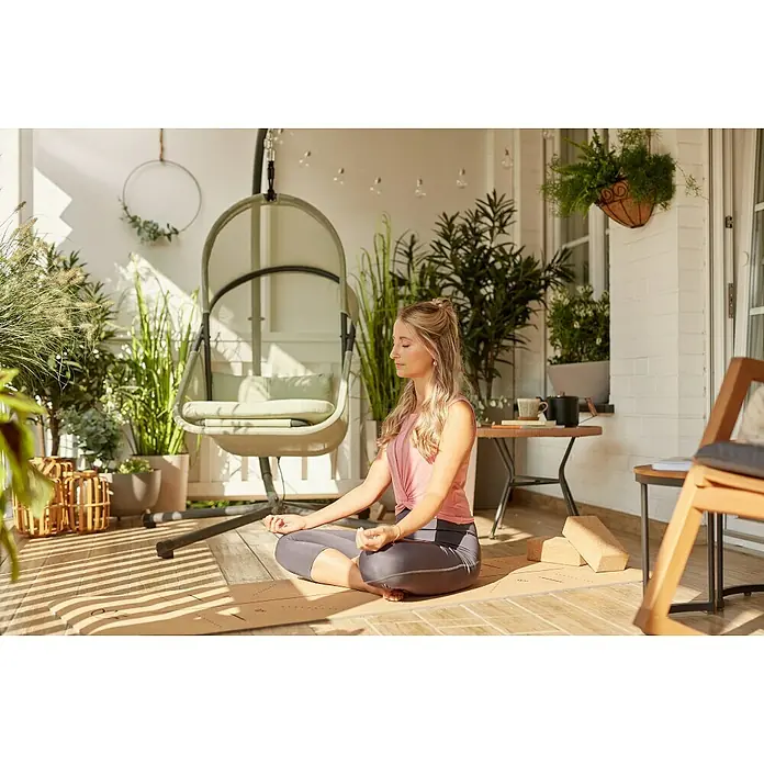 Siena Garden Hängesessel Yobaya mit Schutzhülle - L x B x H: 106 x 123 x 195 cm, Grün - Eine Frau meditiert auf einer Yogamatte, umgeben von Pflanzen, in einem friedlichen Außenbereich mit einem hängenden Stuhl im Hintergrund.