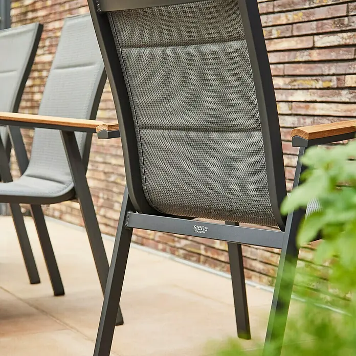 Siena Garden Stapelstuhl Tesoro - 2 Stk., Ranotex®-Gewebe, Stapelbar, Anthrazit, B x T x H: 62,5 x 68 x 98 cm - Nahaufnahme von zwei grauen Gartenstühlen mit Holzarmlehnen vor einer Ziegelwand, die einen stilvollen Außenbereich zeigen.