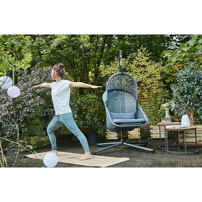 Siena Garden Hängesessel Yobaya mit Schutzhülle - L x B x H: 106 x 123 x 195 cm, Blau - Frau praktiziert Yoga auf einer Matte im Garten neben einem grauen Hängesessel und Beistelltischen.