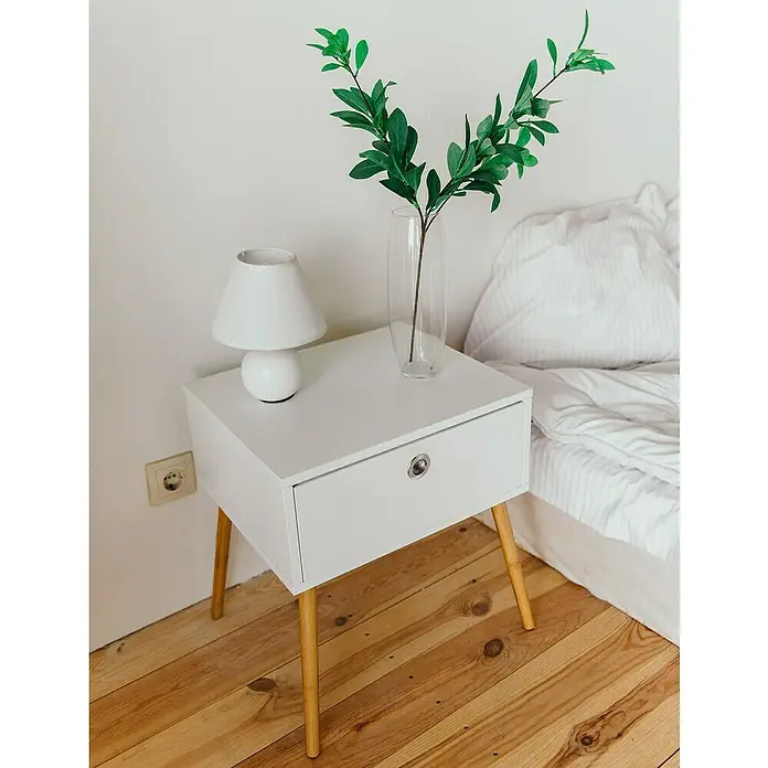 Rei Möbelgriff - Stahl, Nickel - Weißer Nachttisch mit Holzbeinen neben einem Bett, darauf eine Lampe und eine Vase mit Grün.