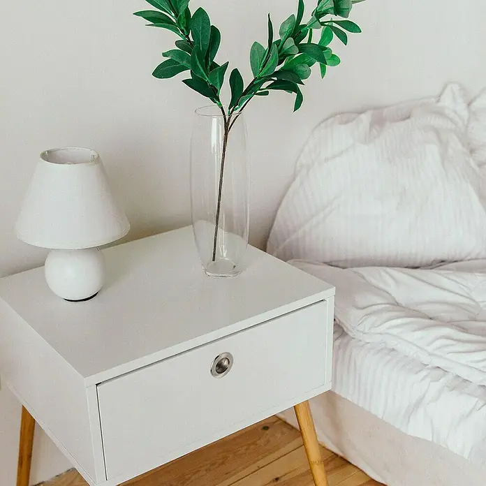 Rei Möbelgriff - Stahl, Nickel - Weißer Nachttisch mit Holzbeinen, auf dem eine Lampe und eine Vase mit grünen Zweigen neben einem weißen Bett stehen.
