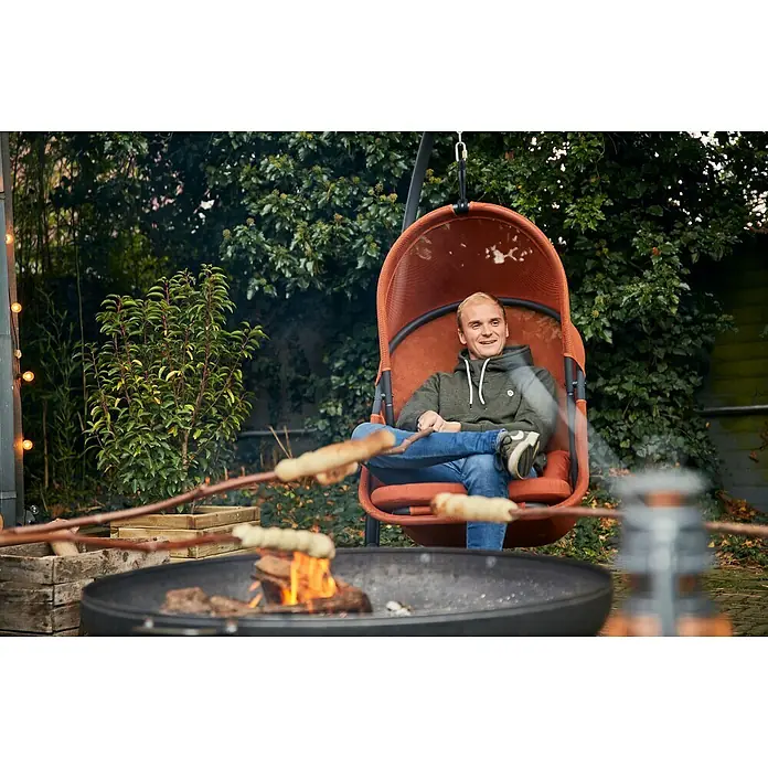 Siena Garden Hängesessel Yobaya mit Schutzhülle - L x B x H: 106 x 123 x 195 cm, Rot - Ein Mann entspannt in einem hängenden Sessel im Garten, während er Stockbrot über einem Feuer grillt. Der Sessel ist ein echter Hingucker.