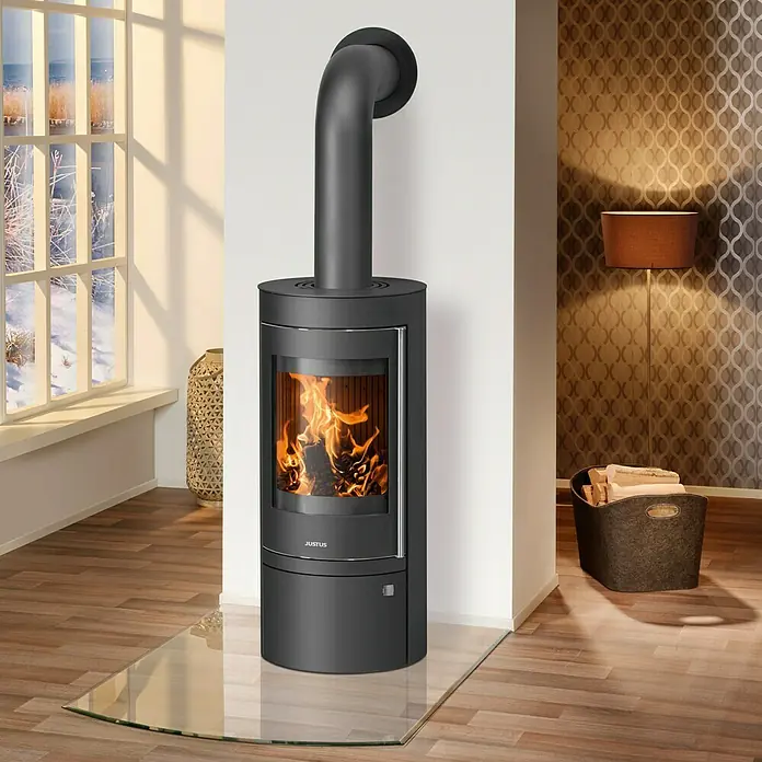 Justus Kaminofen Mino Top 2.0 - 5,5 kW, Raumheizvermögen: 88 m³, Material Verkleidung: Stahl, Schwarz - Kaminofen Justus mit Glasplatte vor einer weißen Wand mit brennendem Feuer und Holzkorb.