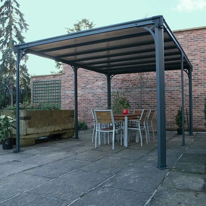 Palram – Canopia Pérgola estándar Milano - An x Pr: 435,9 x 315,5 cm, Color del armazón: Antracita, Aluminio - Una glorieta gris con una mesa de comedor y sillas debajo. La estructura se encuentra en un patio pavimentado con una pared de ladrillos al fondo.