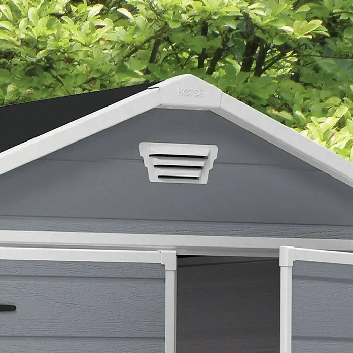 Keter Caseta de jardín Manor 6x8 - Medidas ext. (An x Pr): 185,8 x 236,8 cm, Gris Claro/Blanco, Plástico, Área: 3,9 m² - Primer plano de un cobertizo gris Keter con un tejado a dos aguas blanco y una rejilla de ventilación, rodeado de exuberante vegetación.