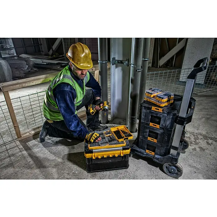 DeWalt T-STAK Transportroller - L x B x H: 59 x 55 x 104,3 cm, Kunststoff, Traglast: 100 kg - Ein Bauarbeiter kniet neben einer DeWalt Werkzeugkiste mit einem gelben Schutzhelm und einer Bohrmaschine in der Hand.
