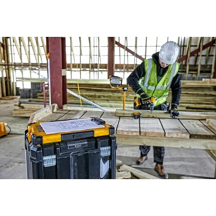 DeWalt T-STAK Portapapeles DWST82732-1 - L x An x Al: 28,5 x 41,3 x 32 cm, Plástico, Luz LED - Caja de herramientas T STAK negra y amarilla con planos en una obra, trabajador al fondo.
