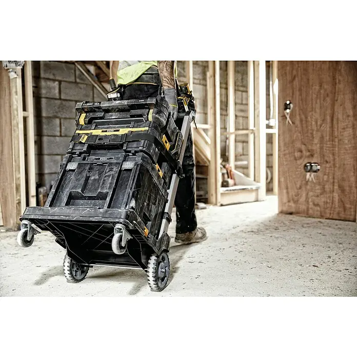 DeWalt T-STAK Transportroller - L x B x H: 59 x 55 x 104,3 cm, Kunststoff, Traglast: 100 kg - Ein Handwerker zieht einen schwarzen Werkzeugkoffer mit gelben Akzenten auf Rollen durch eine Baustelle. Der Werkzeugkoffer ist robust und für den Transport von Werkzeugen ausgelegt.