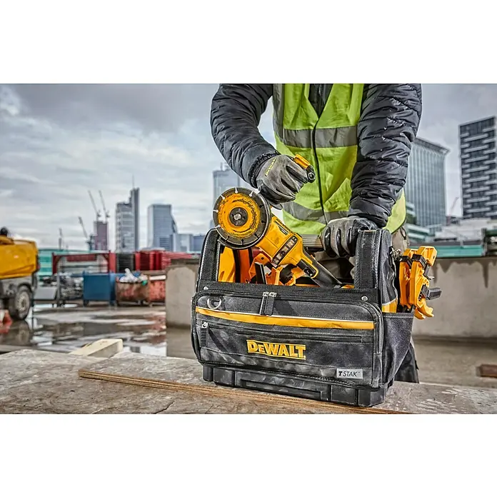 DeWalt T-STAK Bolsa para herramientas - L x An x Al: 25 x 45 x 35 cm, Textil, Capacidad de carga: 30 kg - Una persona coloca una amoladora angular amarilla en una bolsa de herramientas DEWALT TSTAK negra y amarilla en una obra, entre edificios.