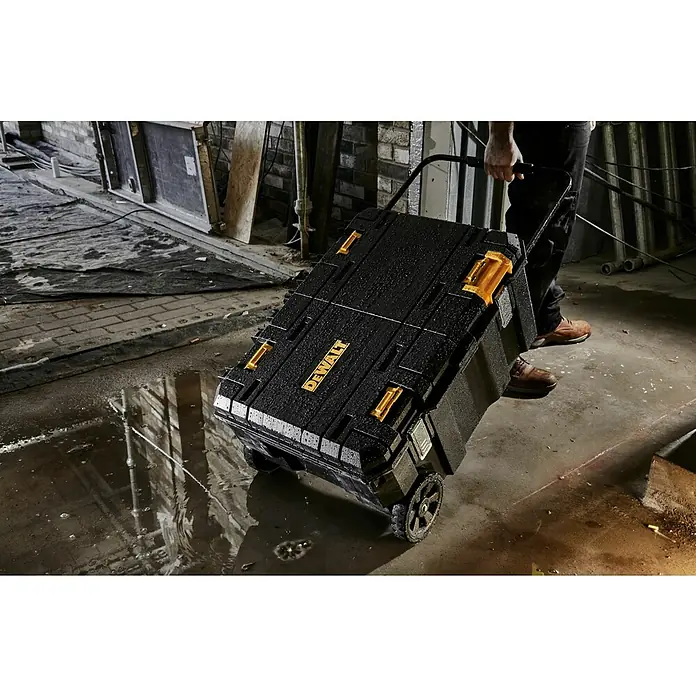 DeWalt T-STAK Caja para herramientas - L x An x Al: 80 x 60 x 43 cm, Plástico, Capacidad de carga: 50 kg - Caja de herramientas DEWALT negra y amarilla con ruedas, tirada por una persona en una zona industrial húmeda, muestra diseño robusto y portátil.