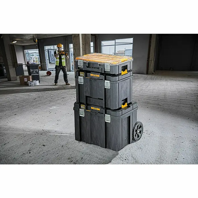 DeWalt Caja para herramientas DWST83346-1 - L x An x Al: 333 x 440 x 302 mm, Plástico, Con cerradura - Pila de cajas de herramientas DeWalt TSTAK negras y amarillas con tapa transparente y ruedas en obra. Trabajador al fondo.