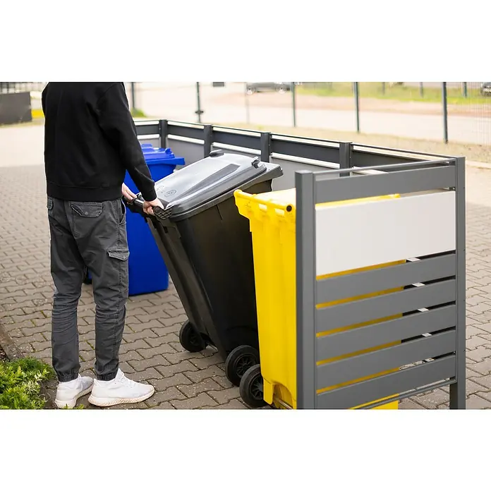 Westmann Mülltonnenbox Verkleidung Planum - 90 x 330 x 120 cm, Anthrazit/Grau - Eine Person schiebt einen schwarzen Mülleimer in einen Mülltonnenständer mit gelbem und blauem Behälter.