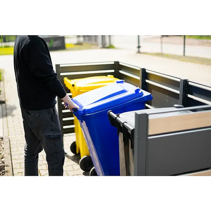 Westmann Mülltonnenbox Verkleidung Planum - 90 x 250 x 120 cm, Anthrazit/Holzoptik - Eine Person schiebt eine blaue Mülltonne mit Rädern in einem Außenbereich mit gelben und grauen Behältern.