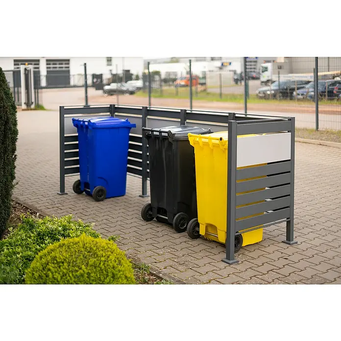 Westmann Mülltonnenbox Verkleidung Planum - 90 x 330 x 120 cm, Anthrazit/Grau - Mülltonnen in blau, schwarz und gelb stehen hinter einer grauen Sichtschutzwand auf einem gepflasterten Bereich im Freien.