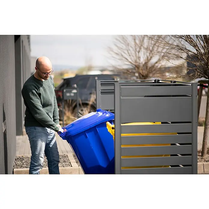 Westmann Mülltonnenbox Verkleidung Planum - 90 x 170 x 150 cm, Anthrazit - Ein Mann mit Brille leert eine blaue Mülltonne in eine graue Mülltonnenbox im Freien, während ein Auto im Hintergrund geparkt ist.