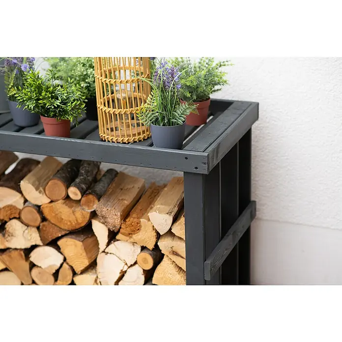 Westmann Kaminholzregal Stephan - 108 x 52 x 74 cm, Holz, Anthrazit - Nahaufnahme eines grauen Holzregals mit Pflanzen und Brennholz vor einer weißen Wand.