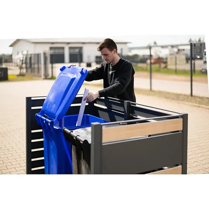 Westmann Mülltonnenbox Verkleidung Planum - 90 x 170 x 120 cm, Anthrazit/Holzoptik - Ein Mann wirft Abfall in einen blauen Mülleimer in einer modernen, grauen Mülltonnenbox mit Holzdetails. Die Box steht auf einem gepflasterten Untergrund.