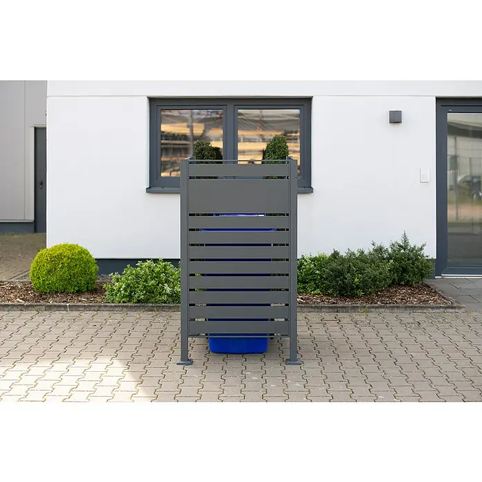 Westmann Mülltonnenbox Verkleidung Planum - 90 x 90 x 150 cm, Anthrazit - Ein grauer Mülltonnenbox-Sichtschutz mit horizontalen Lamellen steht auf einem gepflasterten Hof vor einem weißen Gebäude mit Fenster.