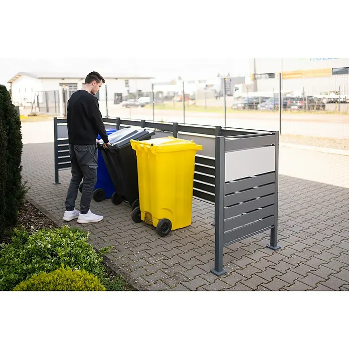 Westmann Mülltonnenbox Verkleidung Planum - 90 x 410 x 120 cm, Anthrazit/Grau - Eine Mülltonnenbox aus grauem Metall mit einem Mann, der eine schwarze Mülltonne hineinschiebt. Daneben stehen eine blaue und eine gelbe Tonne.
