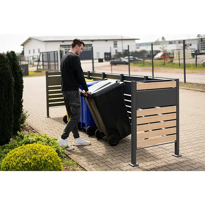 Westmann Mülltonnenbox Verkleidung Planum - 90 x 410 x 120 cm, Anthrazit/Holzoptik - Ein Mann schiebt eine schwarze Mülltonne in eine moderne Mülltonnenbox mit Holzlatten und grauen Paneelen auf einem gepflasterten Weg.