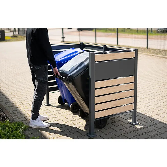 Westmann Mülltonnenbox Verkleidung Planum - 90 x 170 x 120 cm, Anthrazit/Holzoptik - Eine Person stellt eine Mülltonne in eine graue Mülltonnenbox mit Holzverkleidung. Die Box ist für zwei Mülltonnen ausgelegt.